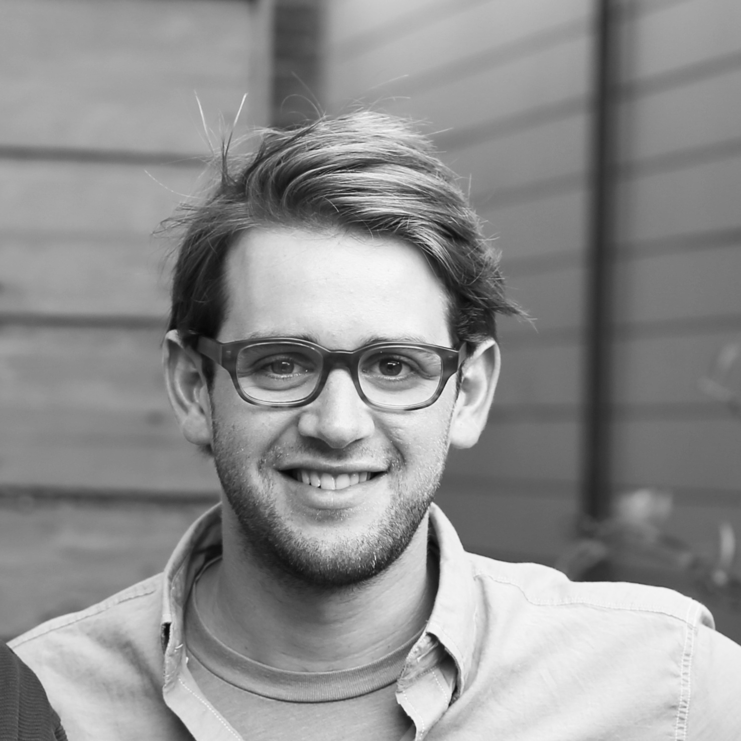 This is a black and white photo of Alexander Fortunado standing in front of a wood paneled building and smiling.