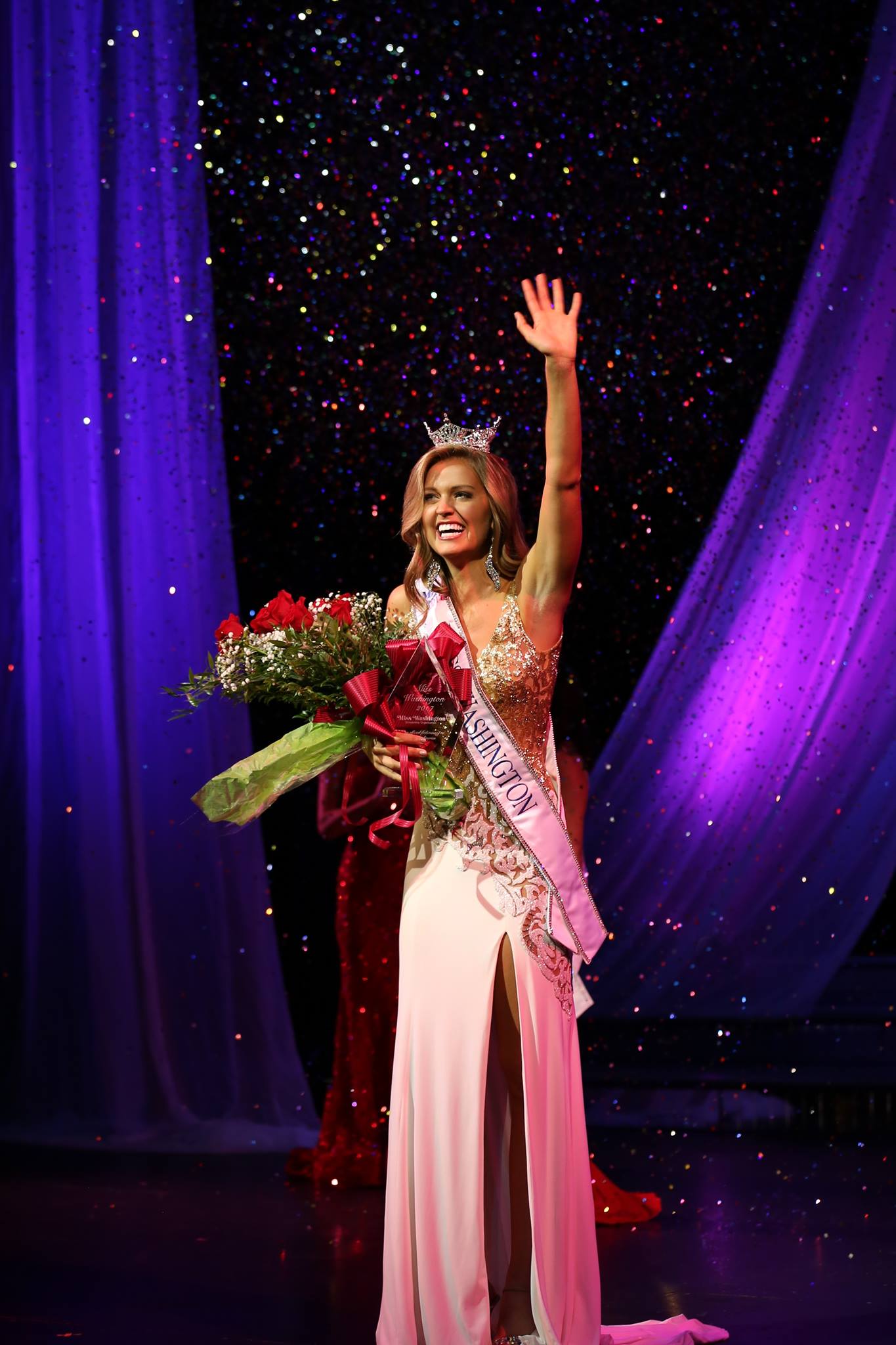Woman in sparking evening gown waves to crowd with one hand, while clutching a large bouquet of roses with the other.