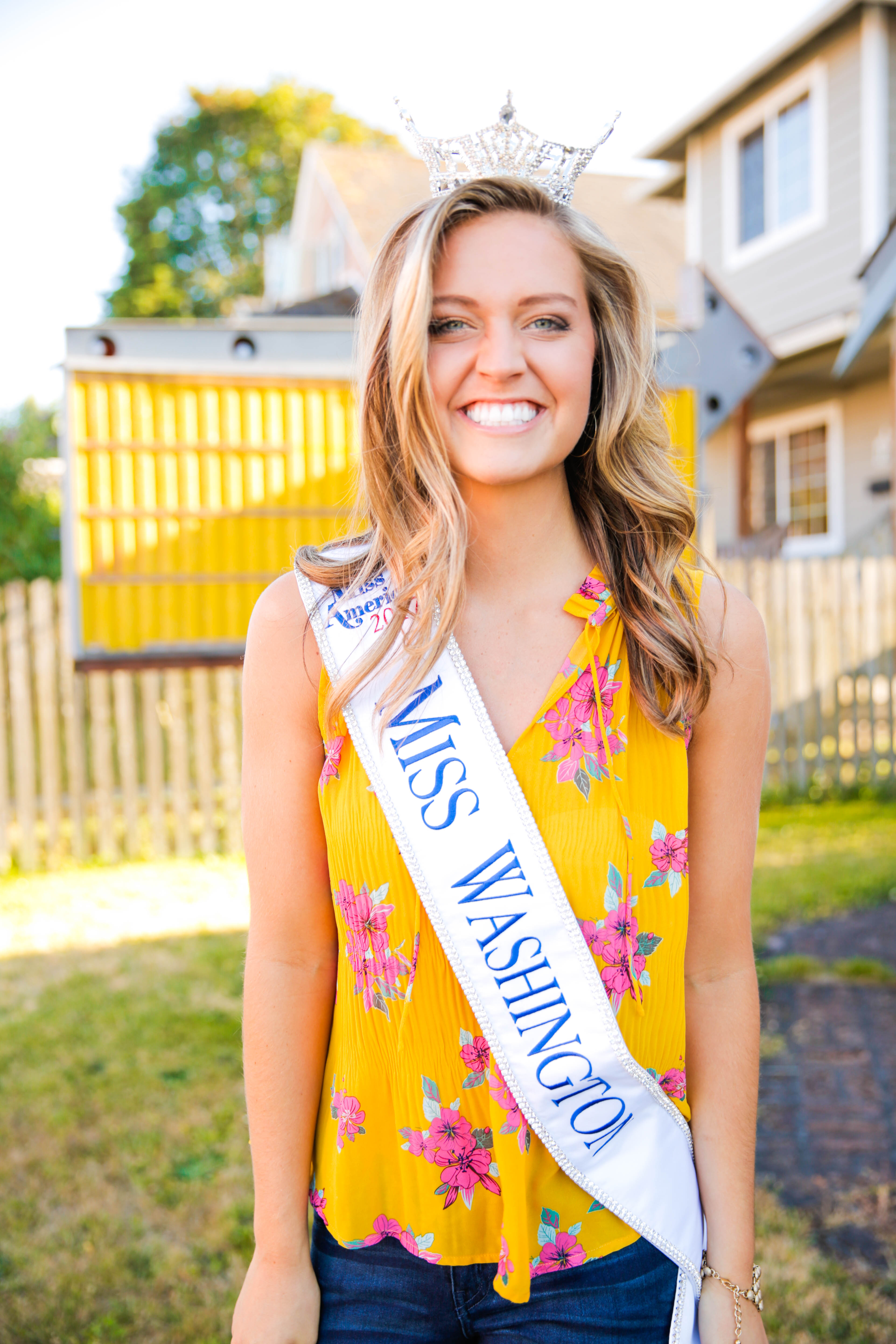 Young woman in her backyard smiles while wearing a tiara and sash from a beauty pageant.