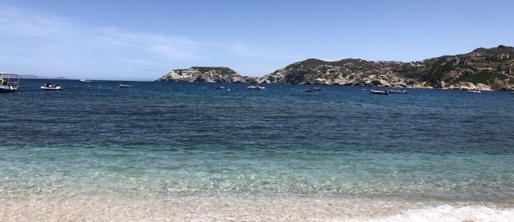 Picture of ocean from beach in Greece. End of a mountain protrudes into the water and ends halfway through the picture. Water begins clear from beach but gets slightly darker about 10 feet in and then increasingly darker after that. Sky is well lit as if it is 3pm in the afternoon. Few clouds present in the distance.