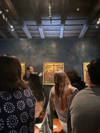 Art gallery. There is someone seemingly explaining a painting. The room is black, dimly lit, and the frame of the painting is gold. The painting is to far to see clearly. The photo is taken from the back of a room filled with about 10 people, some of whose backs block the picture from seeing other paitings around the room.