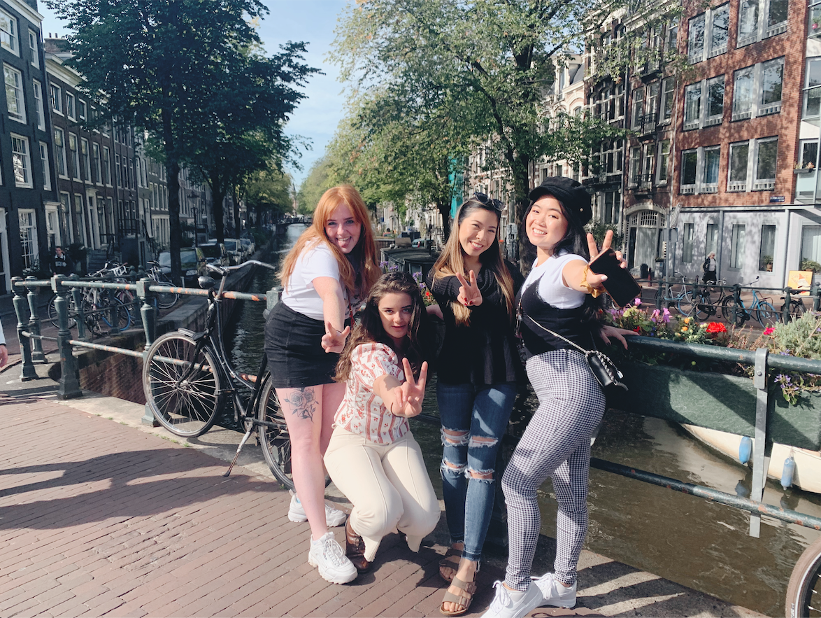 Rosa standing on a bridge and posing with three friends. On the left, a white womxn with red hair wearing a grey shirt and black shorts; her tattoo on her leg is revealed. Kneeling below her is another white womxn with brunette hair. Rosa stands inbetween them and another friend on Rosa's left, who is an Asian womxn. All of them are throwing up the peace sign.