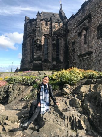 Rosa sitting in front of a castle. The castle is about 3 stories high and is made of grey bricks. Rosa's hair is braided and she is wearing black jeans and a black leather jacket along with a blue plaid scarf.