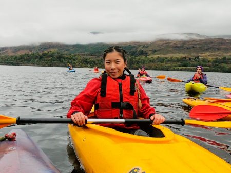 Rosa stopped on her kayak. Wearing a red life vest and a matching red wool sweater. Her hair is braided and she is smiling. Kayakers are featured in the background and the land is about a mile in the distance.