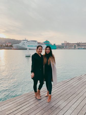 Rosa standing with a friend on a pier with a cruise boat in the background and the sun setting behind a hill with clouds overhead. Rosa is wearing black pants and a black shirt and brown boots.