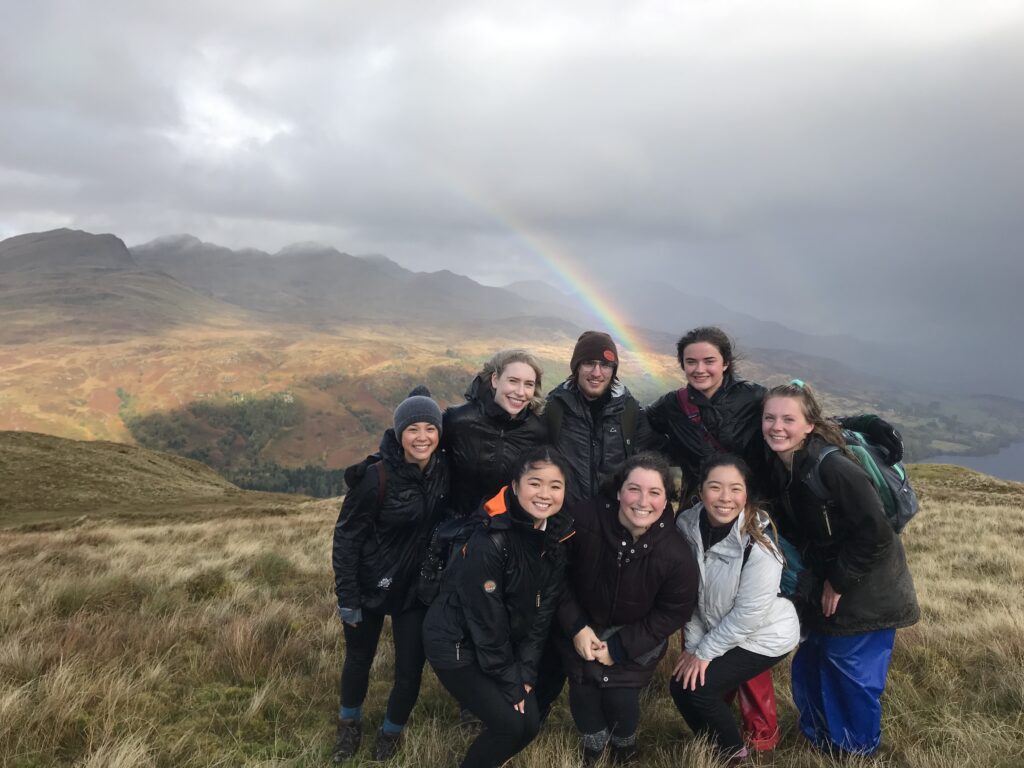 Rosa with friends on top of a mountain of beige/green grass fields. There are grey storm clouds in the background and rain in the distance. Sun shines through a hole in the clouds and a rainbow arches out behind the group. The group is primarily wearing black overcoats.