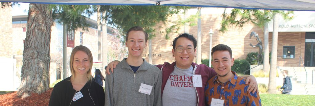 4 student workers standing at a table during study abroad fair. They are hugging each other, smiling, and looking at the camera.