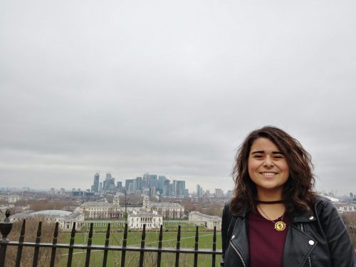 Talia with the London skyline in the background. She is standing in front of a black fence with a large green lawn between her and the skyline.
