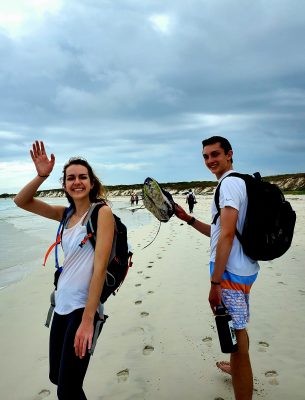 Students look back and waive at the camera while walking on the beach