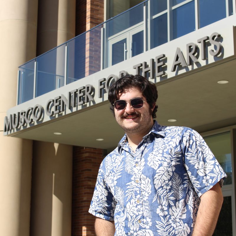 Young man with short dark hair, mustache and sunglasses standing in front of Musco Center.
