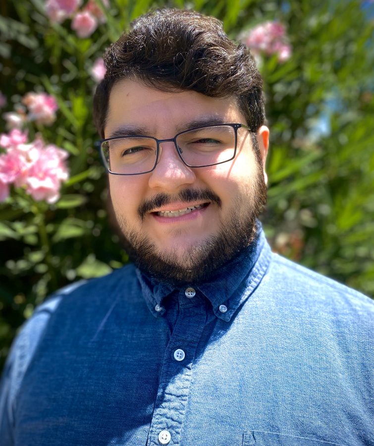 Smiling young man with dark short hair, beard, and dark-rimmed glasses in a blue chambray shirt.