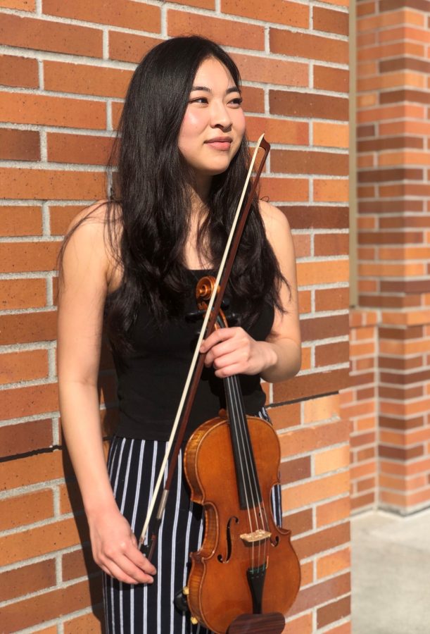 Smiling young woman with long dark hair wearing black and white striped pants and black tank top, holding a violin.