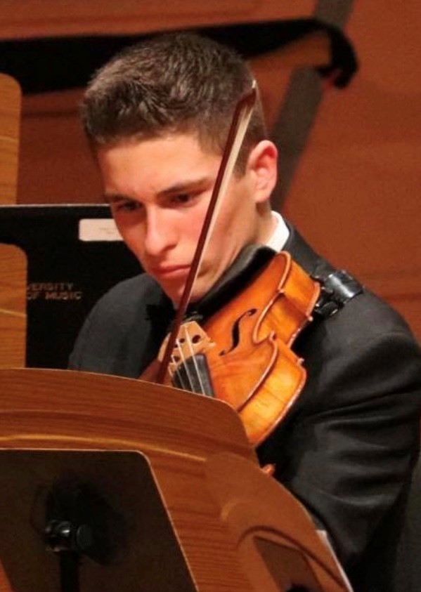 Young man with short dark hair in a tuxedo playing the viola and intensely focused on sheet music in front of him.