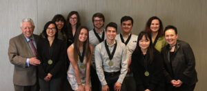 Group of smiling young people wearing medals and two elderly people standing on each side of the group.