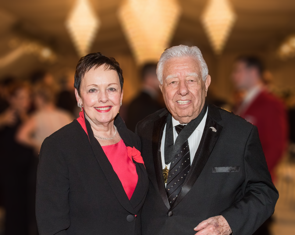 Smiling older woman with short black hair in a black suit with red blouse; smiling older man with white hair in black tuxedo