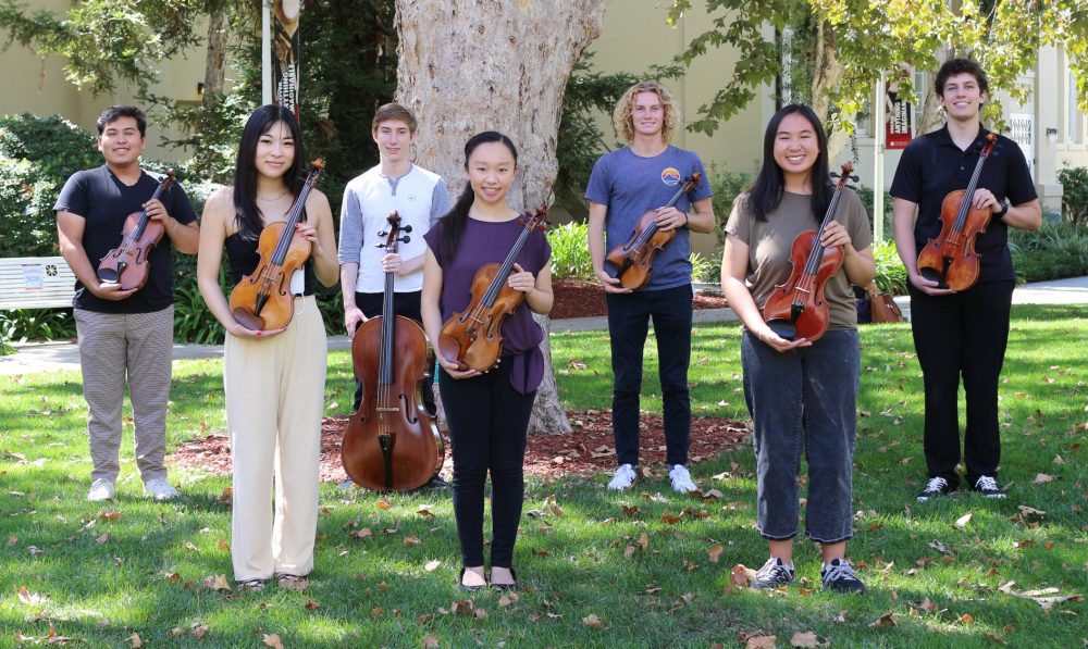 Seven string students lined up on a lawn holding their instruments.
