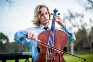 Young man with shoulder-length blonde hair wearing a light blue dress shirt is seated on a park bench playing a cello.