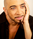 African-American man with shaved head and narrow goatee sits with hand on side of face