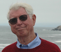 white-haired man with sunglasses wearing red sweater over blue button-down shirt with ocean in the background
