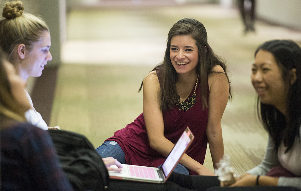 Students sit on floor with one laptop talking.