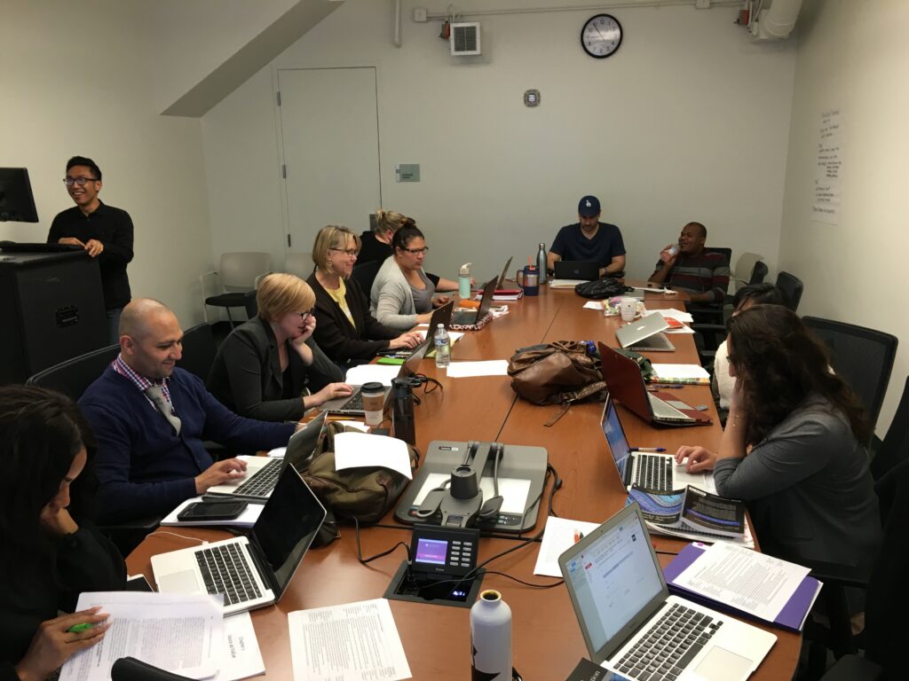 Man stands at a computer, typing, while others sit at conference table with laptops.