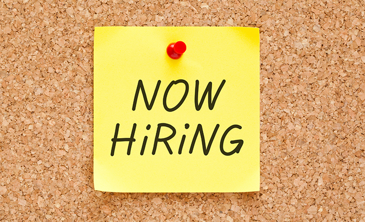 Now Hiring written on a yellow post-it pinned to a cork board