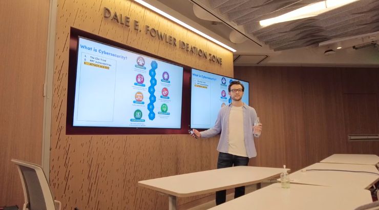 A young man with dark-framed glasses, standing in front of his presentation. He is holding his phone in one hand, and a water bottle in the other.
