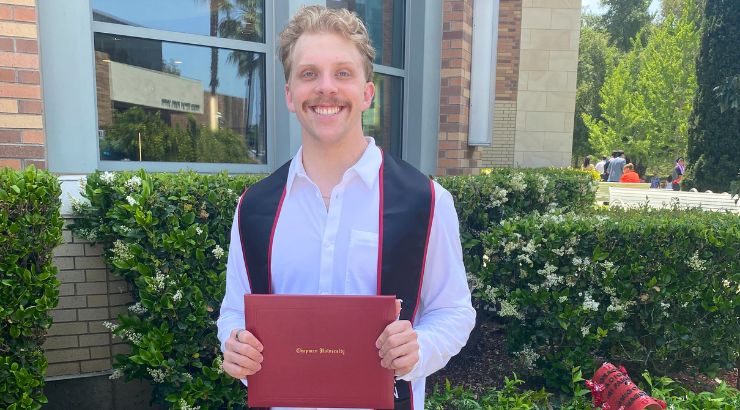 Jackson Thomas standing in front green brushes wearing a graduation sash and holding his degree.