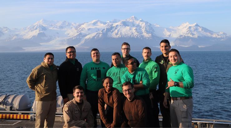 A team of US Navy standing posing together with a snowy mountain in the background.