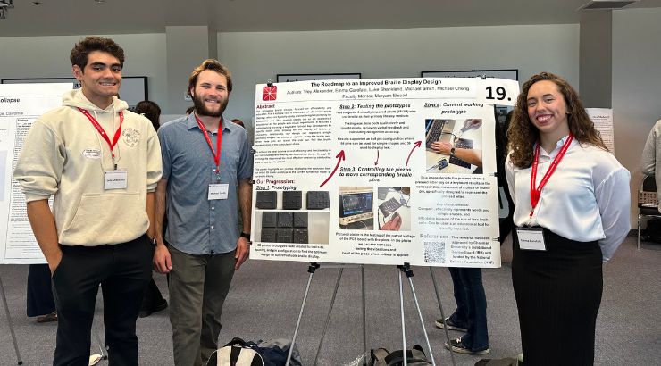 A group of three engineering students proudly stand in front of their research presentation. 