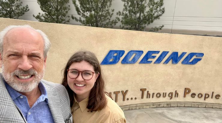 A young engineering students is posing for a selfie with her father on the left-hand side. On the right-hand side is the sign "Boeing".