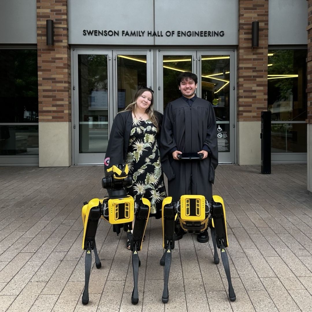 Ethan Tapia (right) with Kalin Richardson (left), dressed in black commencement gowns, posing with the SPOT dogs in front of Swenson Hall