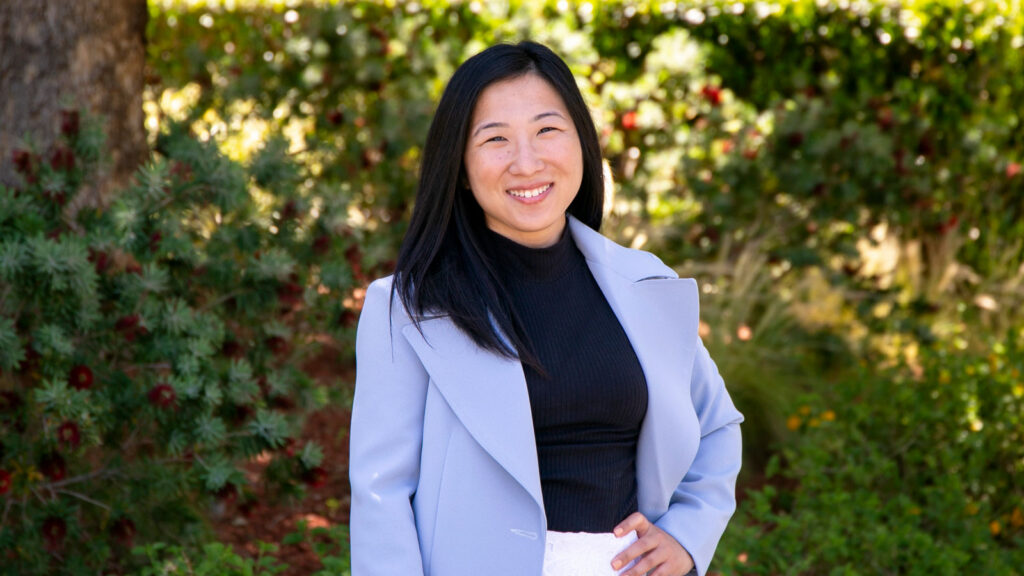 Caroline Nam in a light blue jacket standing in front of green foliage