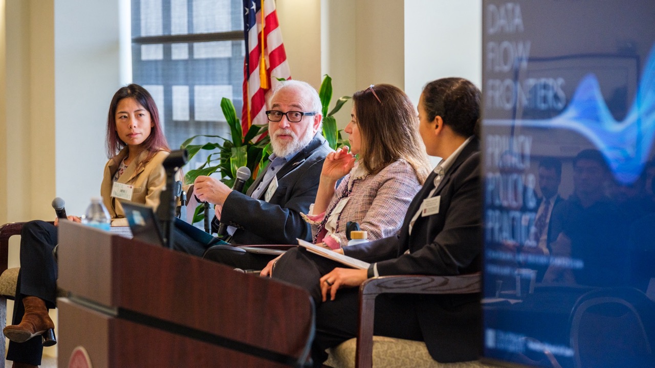 A group of four panelists pictured near a screen displaying the poster for this event.