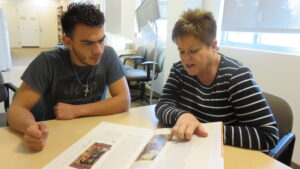 A seated adult woman, right, shows a seated young adult male student, left, something in a book.