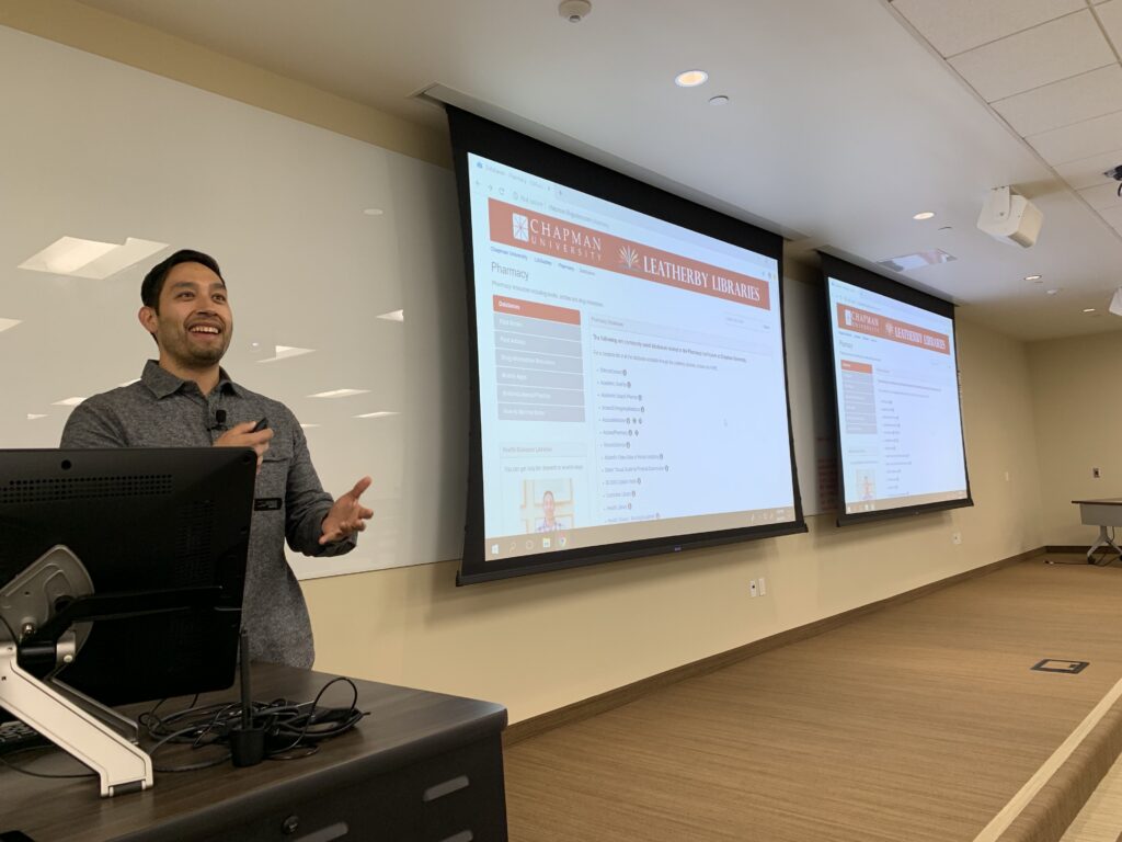 A young male librarian stands at a computer podium with two projector screens behind him displaying the library website.