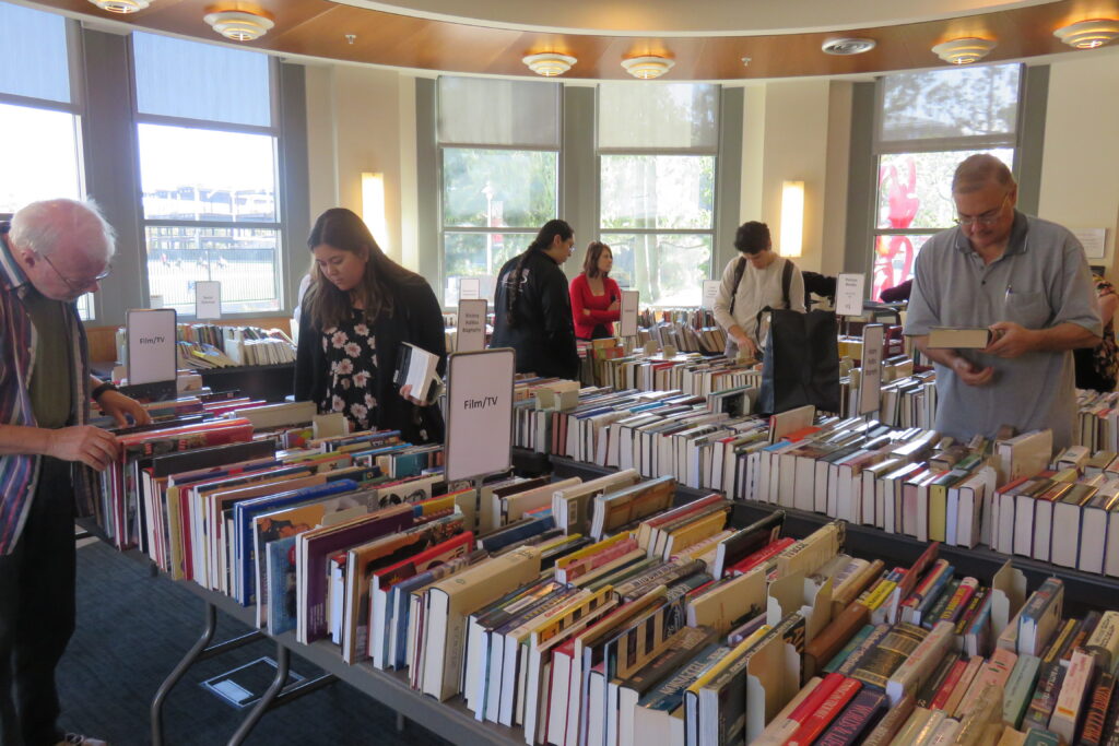Several adults, some in the foreground and some in the back, look through tables of books, organized by genre.