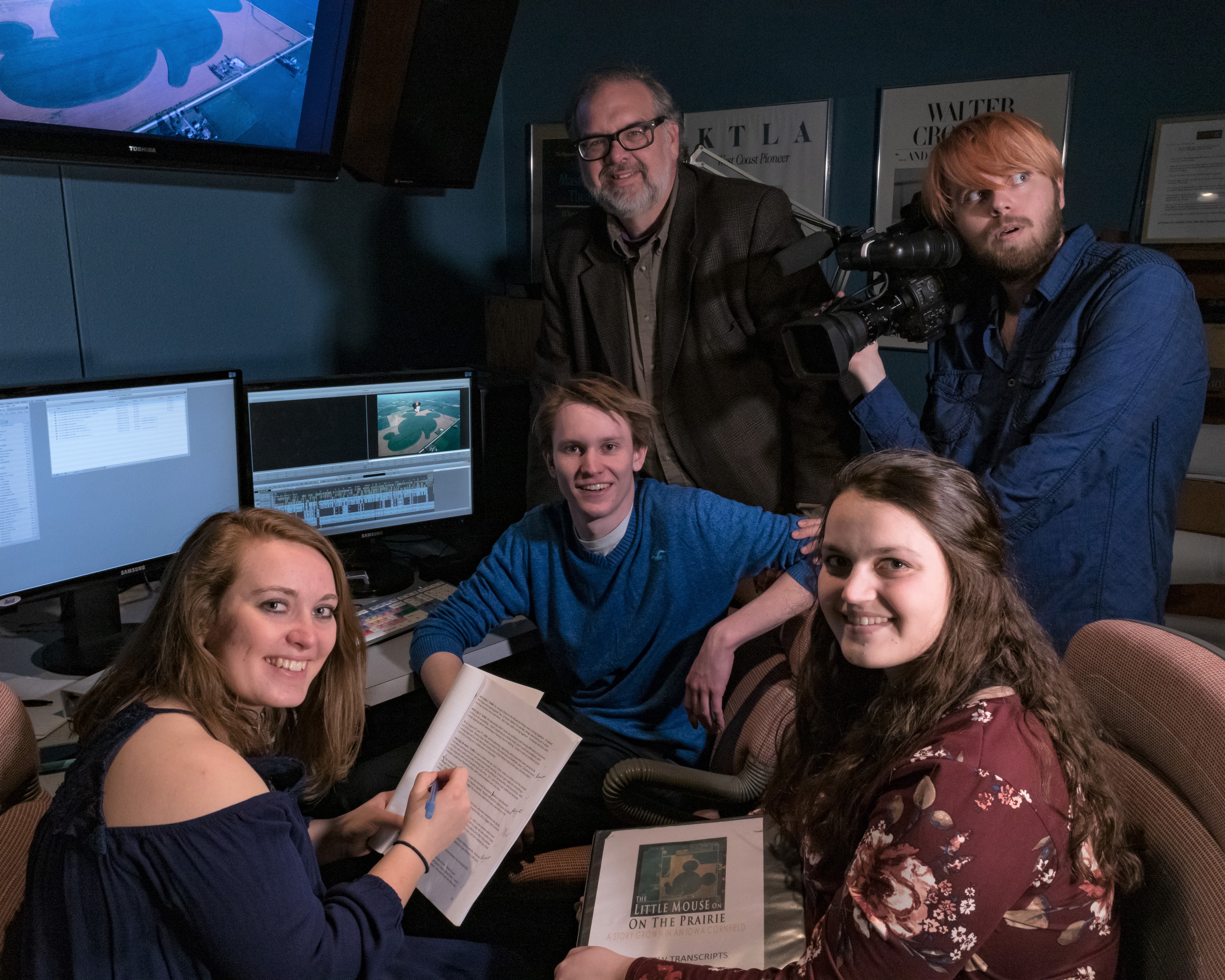 Group of four students and one professor sitting and standing around a computer, with scripts open on their laps.