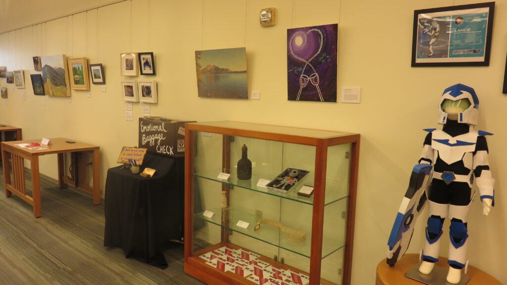 Several different art pieces lined up on a wall. Some are paintings or photographs hung on the wall, some are displayed in a display case, and one sculpture on the far right.