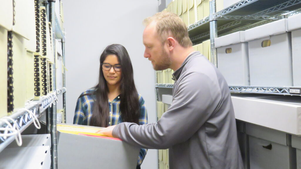 A young woman (r.) and a young man (l.) look at documents in a bankers box, in between shelves of boxes.