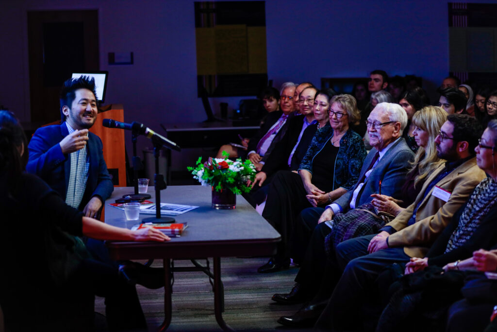 A man seated at a table energetically engages with an audience.