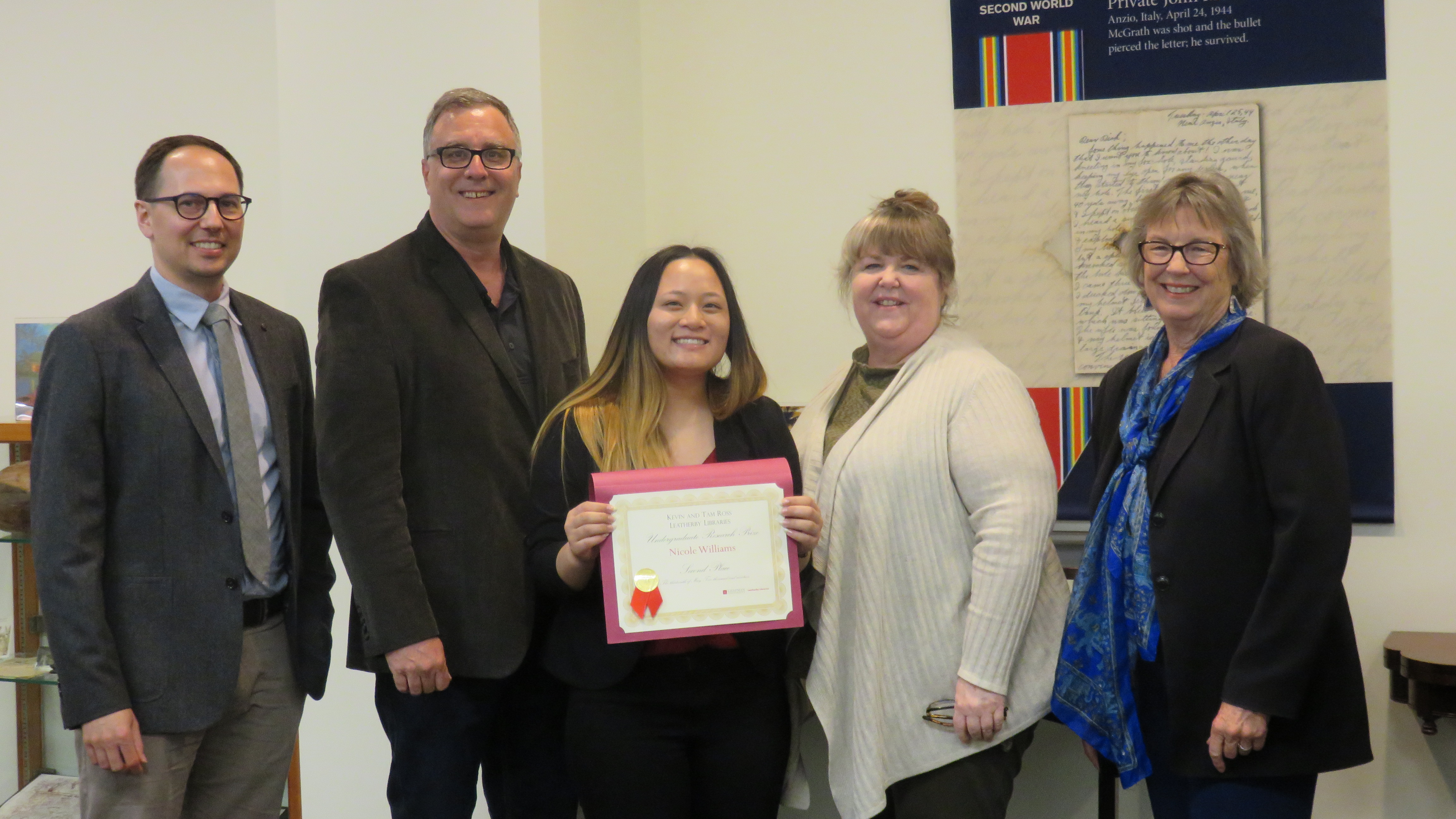 Two men stand on the left, with three women on the right. The young woman in the middle holds a certificate.