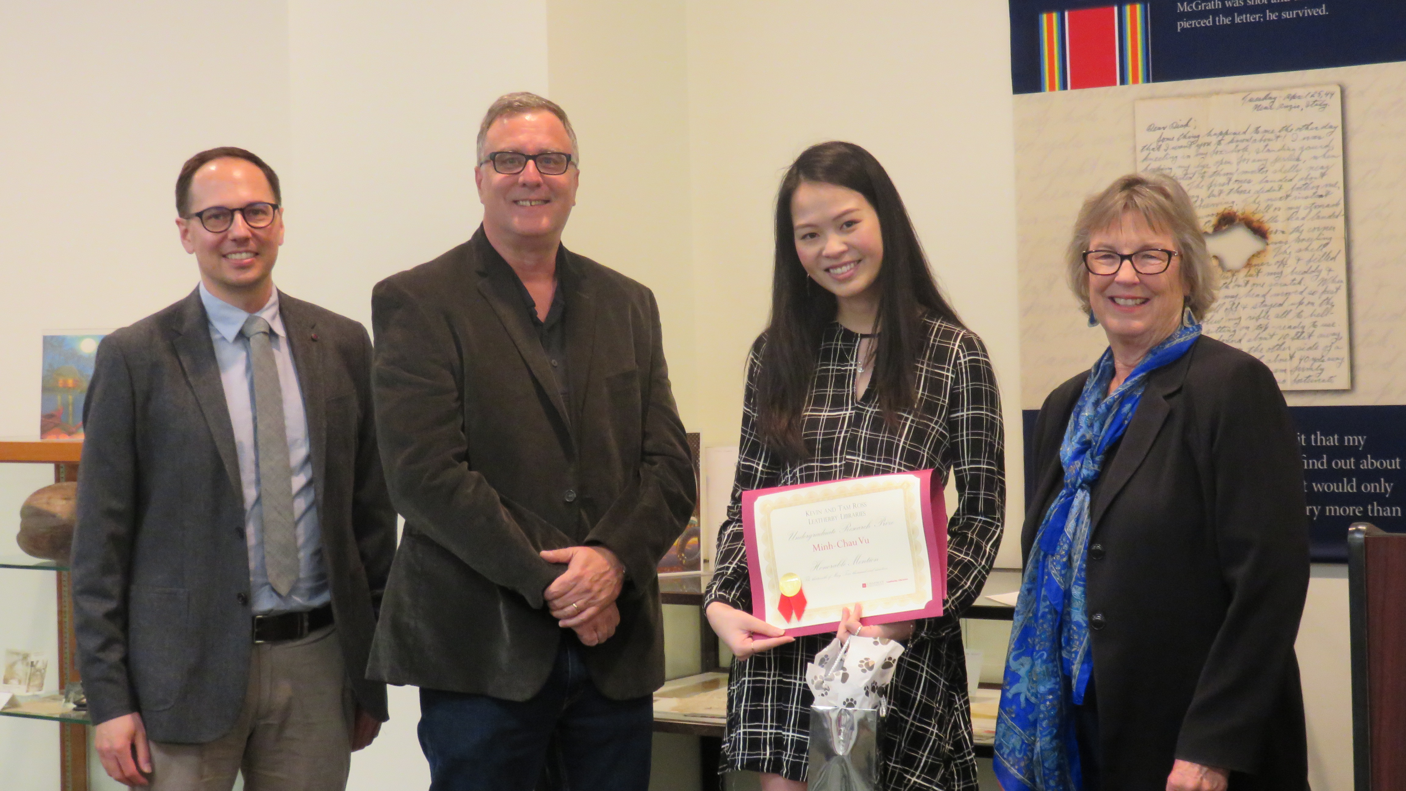 Two men stand on the left, and two women on right. The young woman in the middle holds a red certificate.