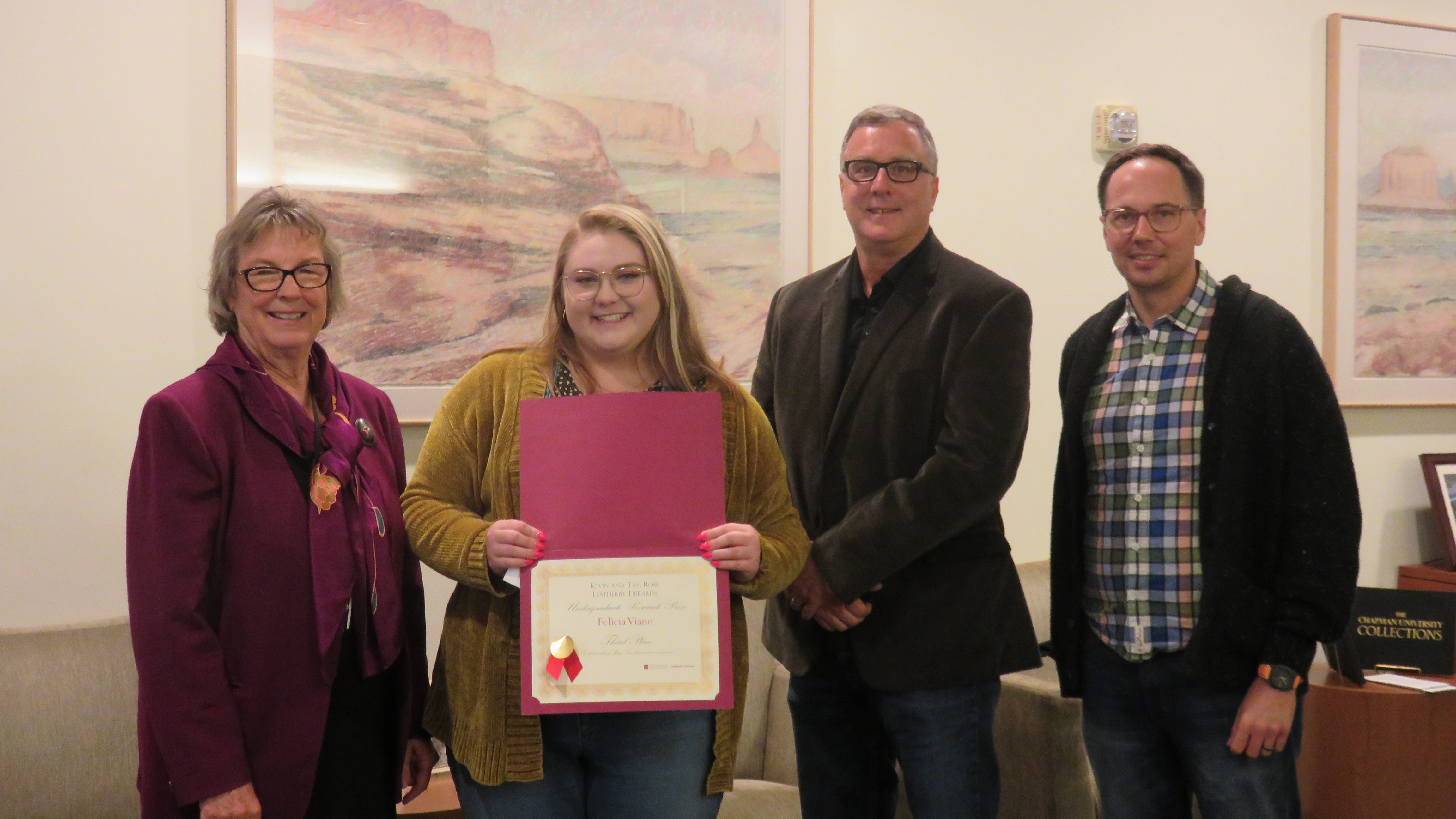 Two women stand on the left, and two men on the right. The young woman second from the left is holding a red certificate.