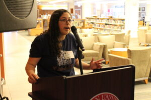 A young woman in a t-shirt stands behind a podium, speaking into a microphone.