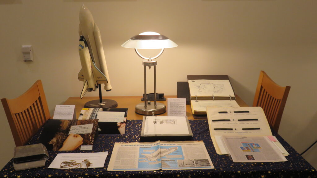 Multiple materials, some printed, some photographs, and one model of the Challenger shuttle, displayed on a table with a lamp in the background.