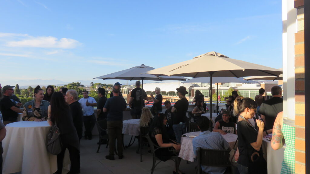 A crowd of people sit and stand on a patio, with a blue sky and mountains in the background.