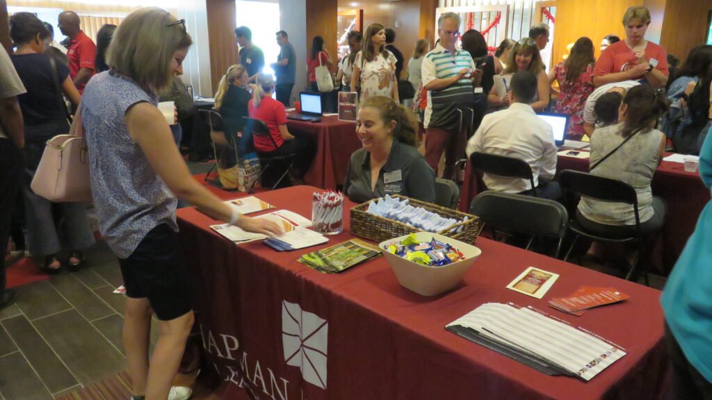 A woman sits at a table with papers and giveaways on it. A woman approaches her, and a crowd is in the background.