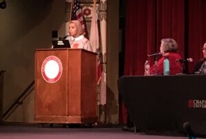 A woman stands at a podium while another woman sits at a table looking at her