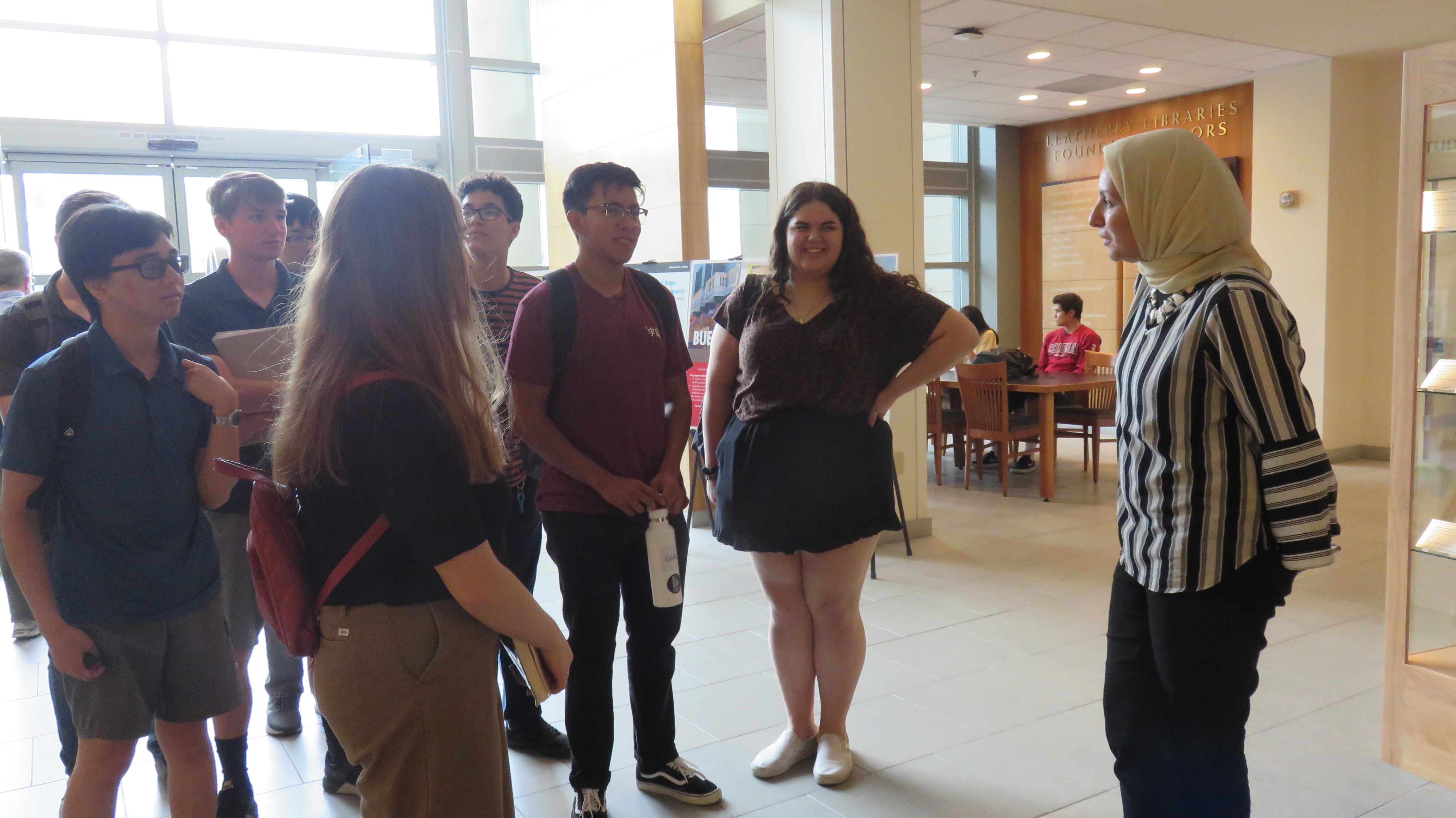 A woman in a headscarf stands to the right of the frame, and a group of high school-aged boys and girls stand to the left.